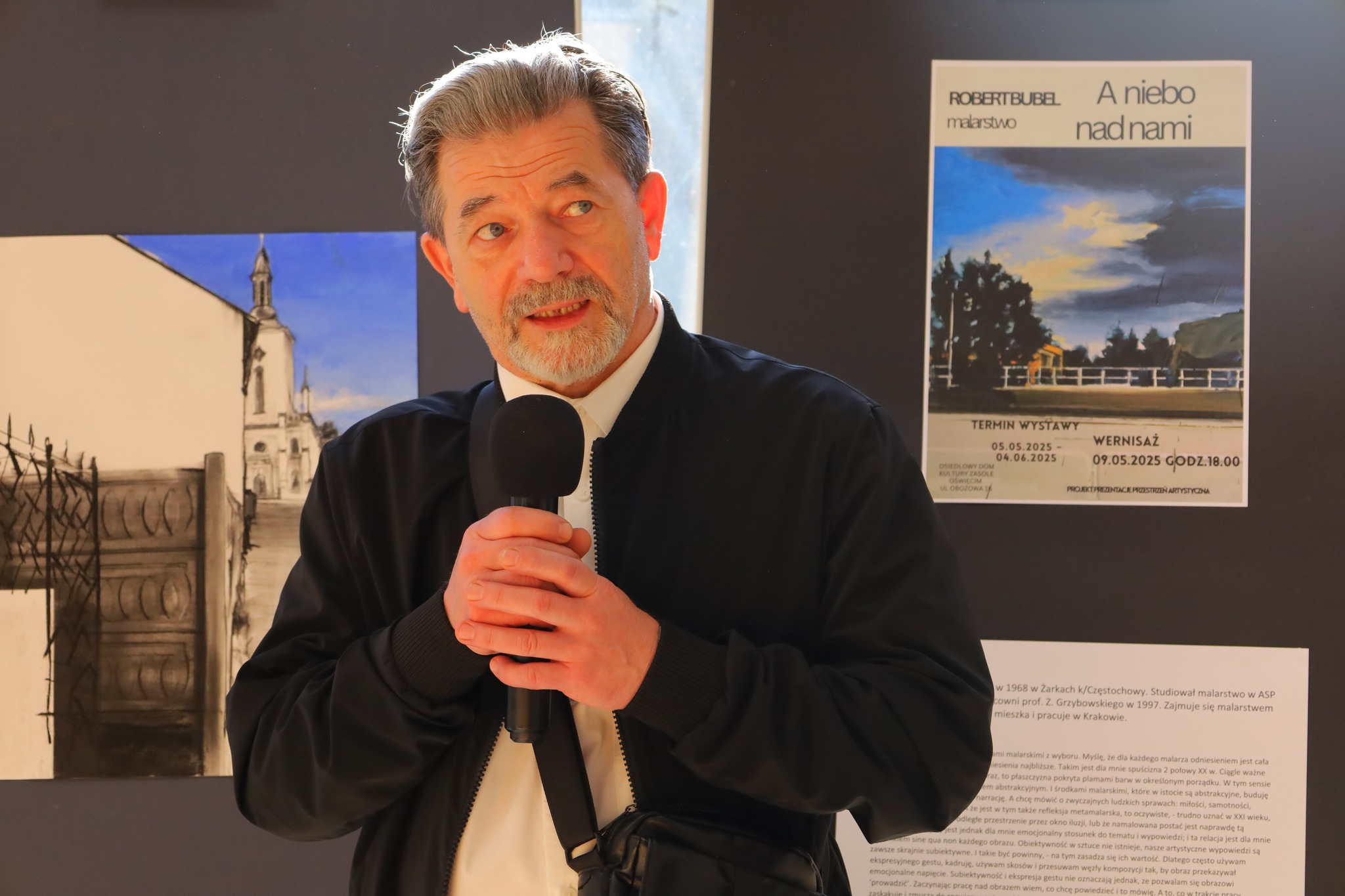 Malarz Robert Bubel podczas swojego wernisażu na tle obrazu z błękitnym niebem